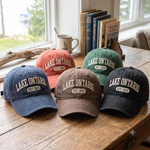 May include: Five baseball caps in blue, orange, brown, green, and black. Each cap features the embroidered text "LAKE ONTARIO" in an arched font above a smaller rectangular patch that reads "EST. 1615" in white lettering on a dark background.