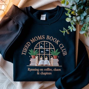 Może przedstawiać: Granatowa bluza z haftowanym wzorem. Wzór zawiera napis "Tired Moms Book Club" wokół łukowatego okna z książkami, świecami i roślinami. Poniżej okna znajduje się napis "Running on coffee, chaos & chapters".
