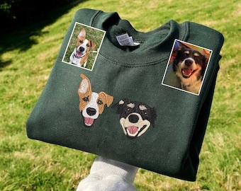 Puede incluir: Sudadera verde oscuro con retratos de perros bordados. Dos caras de perro bordadas están en la parte delantera, con dos fotos de perro adicionales en las esquinas superiores. La sudadera se sostiene contra un fondo de hierba verde y árboles.