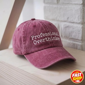 May include: A maroon baseball cap with a distressed, washed-out finish. The front of the cap features the white embroidered text "Professional Overthinker" in a clean, sans-serif font. The cap has a curved brim and a structured crown.