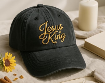 May include: A dark gray baseball cap with the embroidered text "Jesus is King" in gold script. The cap has a curved brim and a button on top. The image also includes a candle, a book, and a sunflower.