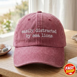 May include: A maroon baseball cap with the embroidered text "easily distracted by sea lions" in white. The cap is made of a soft, textured fabric and has a curved peak. The hat is on a wooden surface.