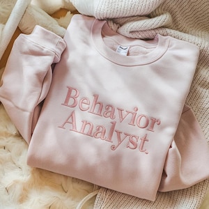 May include: A pale pink sweatshirt featuring the embroidered words "Behavior Analyst" in a complementary pink hue. The garment is neatly folded, placed on a textured surface, implying a comfortable, relaxed style.