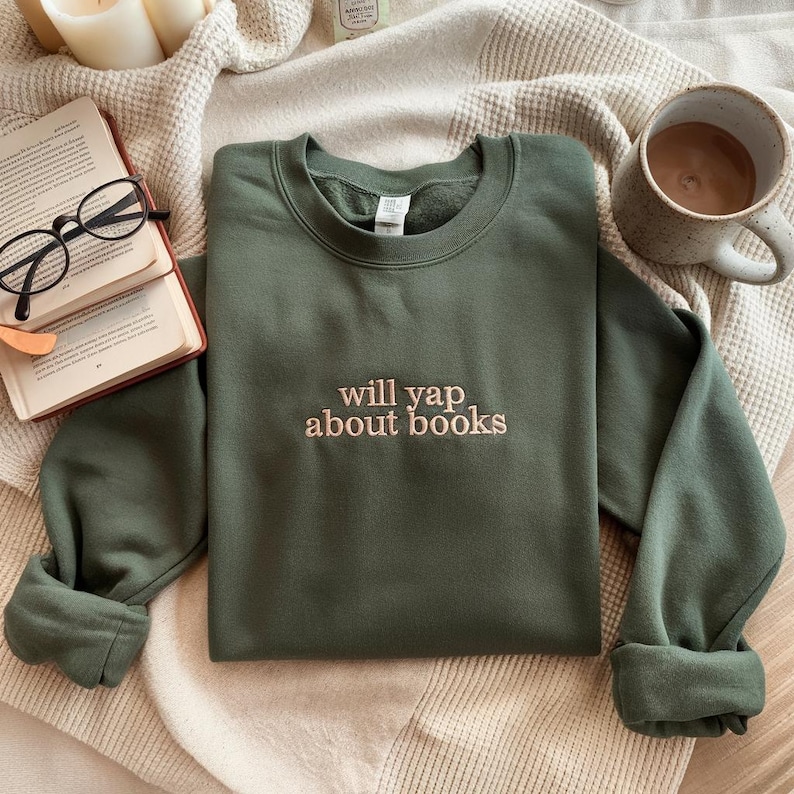 May include: An olive green sweatshirt with the embroidered text "will yap about books" in a light brown color. The sweatshirt is folded on a beige blanket, with a book, glasses, and a mug of coffee nearby.