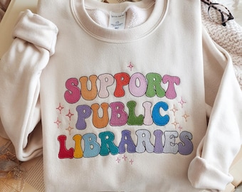 May include: Cream-colored sweatshirt with the embroidered text "SUPPORT PUBLIC LIBRARIES" in a retro, colorful font. The design includes stars and a rainbow of colors. The sweatshirt is folded on top of open books.