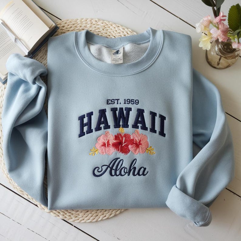 May include: Light blue sweatshirt with embroidered text "EST. 1959 HAWAII Aloha" and three pink hibiscus flowers.