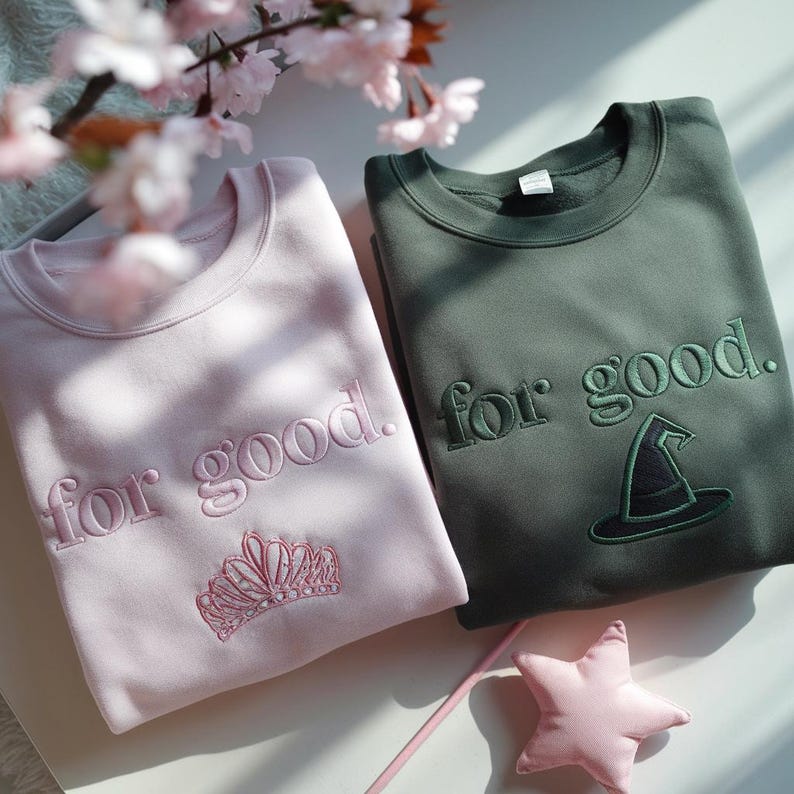 K&ouml;nnte beinhalten: Zwei Sweatshirts, eines hellrosa und eines olivgr&uuml;n, mit dem Schriftzug "for good" bestickt. Das rosa Sweatshirt zeigt eine Krone, das gr&uuml;ne einen Hexenhut. Ein rosa sternf&ouml;rmiger Zauberstab liegt daneben.