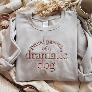 May include: A light gray sweatshirt with the embroidered text "proud parent of a dramatic dog" in a brown color. The sweatshirt has a crew neck and long sleeves. The image also shows a leather belt, a mug, and dried flowers.