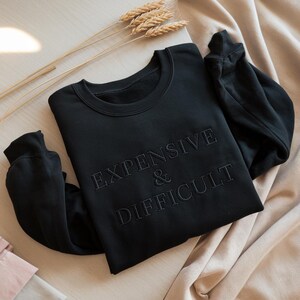 Puede incluir: Sudadera negra de cuello redondo con el texto bordado "EXPENSIVE & DIFFICULT". La sudadera está hecha de un material suave y está sobre una superficie beige. Espigas de trigo secas están colocadas en la superficie.
