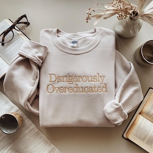 May include: A light beige sweatshirt with the embroidered text "Dangerously Overeducated" in a gold color. The sweatshirt is displayed on a neutral surface with a book, glasses, and decorative items.