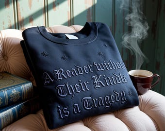 Può includere: Felpa blu navy con ricamo: "A Reader Without Their Kindle is a Tragedy." La felpa ha dettagli a stella ed è piegata su una sedia trapuntata. Una tazza di caffè e libri sono anche nell'immagine.