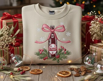 May include: Beige sweatshirt with an embroidered Dr. Pepper bottle design, wrapped with a pink bow and snowflakes. The design includes berries and greenery, perfect for the holiday season. Christmas-themed apparel.