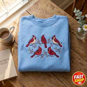 Puede incluir: Una sudadera azul claro de cuello redondo con un diseño bordado de cinco cardenales rojos posados sobre finas ramas marrones con pequeñas bayas rojas. El bordado está centrado en la parte del pecho de la prenda.