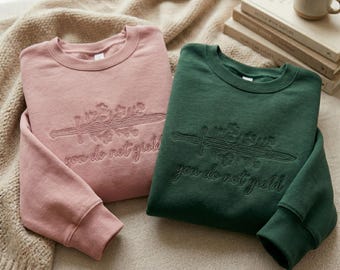 May include: A dusty pink sweatshirt and a dark forest green sweatshirt, both featuring long sleeves and a crew neckline. Each garment displays an embroidered design of a sword intertwined with floral elements and the text you do not yield.
