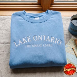 May include: A light blue crewneck sweatshirt featuring white embroidered text that reads LAKE ONTARIO and THE GREAT LAKES in a curved font across the chest. The garment has a ribbed collar and long sleeves, showcasing a clean, simple design.