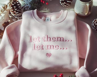Op de afbeelding: Lichtroze sweatshirt met de geborduurde tekst "let them... let me..." en een hartje. Het kledingstuk wordt getoond op een witte ondergrond, omringd door feestelijke decoraties zoals dennenappels en rode bessen.