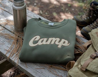Puede incluir: Una sudadera verde oliva con la palabra "Camp" en cursiva color crema. Un termo plateado y una mochila caqui también son visibles sobre una mesa de picnic de madera. Botas de senderismo en el fondo.