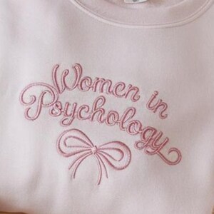 May include: A pink sweatshirt with the text "Women in Psychology" embroidered in pink script with a pink ribbon below the text.