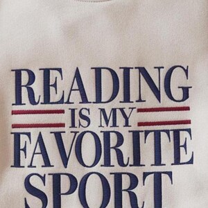 K&ouml;nnte beinhalten: Cremefarbenes Sweatshirt mit dem gestickten Text "READING IS MY FAVORITE SPORT" in Marineblau, mit roten horizontalen Linien. Das Sweatshirt hat einen Rundhalsausschnitt und ein weiches, strukturiertes Aussehen.