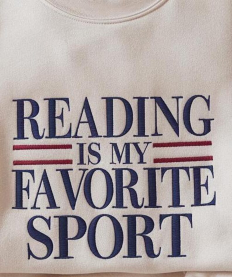 K&ouml;nnte beinhalten: Cremefarbenes Sweatshirt mit dem gestickten Text "READING IS MY FAVORITE SPORT" in Marineblau, mit roten horizontalen Linien. Das Sweatshirt hat einen Rundhalsausschnitt und ein weiches, strukturiertes Aussehen.
