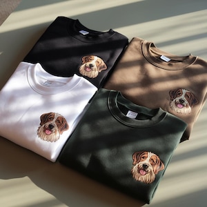 May include: Four sweatshirts in different colors, each featuring an embroidered image of a brown and white dog with a happy expression. The sweatshirts are black, brown, white, and green.