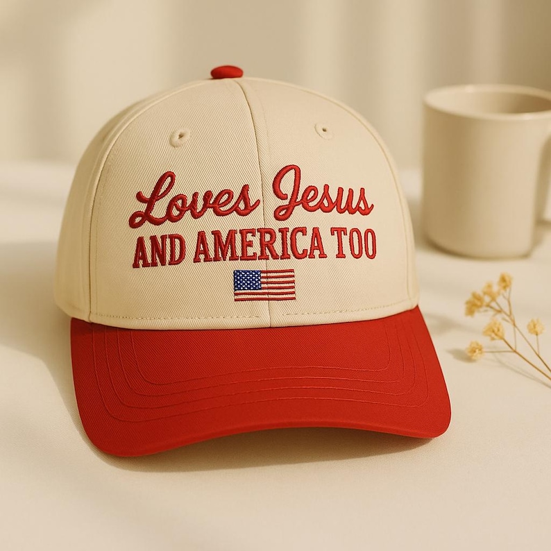 May include: A baseball cap with a cream-coloured crown and a red brim. The cap features the embroidered text "Loves Jesus AND AMERICA TOO" in red, along with a small American flag. The cap has a red button on top.