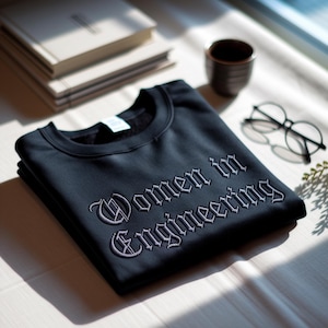 May include: A black sweatshirt with the embroidered text "Women in Engineering" in a gothic font. The sweatshirt is folded neatly on a white surface, with a stack of books, a small cup, and eyeglasses in the background.