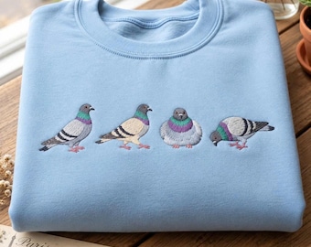 May include: A light blue crewneck sweatshirt featuring an embroidered design of four pigeons in various poses. The pigeons are depicted with grey, white, and iridescent purple and green neck feathers, showcasing detailed stitching across the chest area.