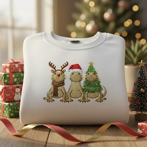 May include: White crewneck sweatshirt featuring embroidered Christmas-themed bearded dragons. One dragon wears reindeer antlers and a scarf, another a Santa hat, and the third is a Christmas tree. Festive holiday apparel.