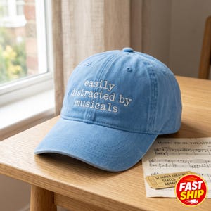 May include: A light blue baseball cap with the embroidered text "easily distracted by musicals." The cap is made of a soft fabric and features a curved brim. A musical score and a theater ticket are on the table.
