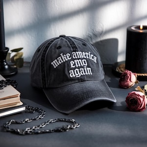 May include: A dark gray baseball cap with the phrase "make america emo again" embroidered in white. The hat is sitting on a dark surface with a black candle and roses in the background.