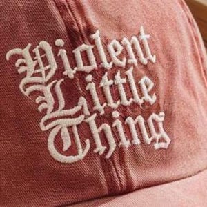 May include: A faded red baseball cap featuring the text Violent Little Thing embroidered in white thread using a gothic-style font. The fabric has a distressed, washed-out appearance with visible stitching details across the crown panels.