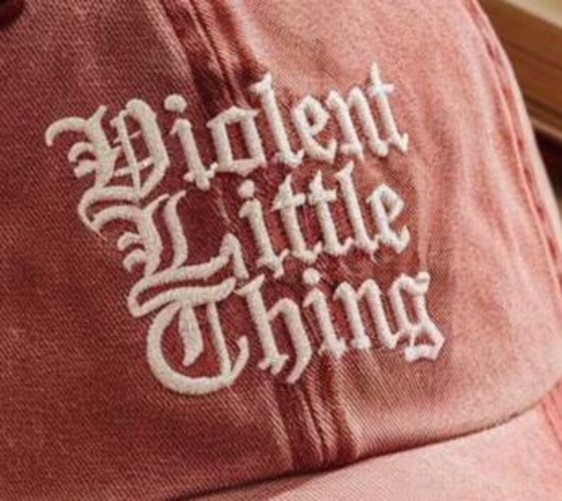 May include: A faded red baseball cap featuring the text Violent Little Thing embroidered in white thread using a gothic-style font. The fabric has a distressed, washed-out appearance with visible stitching details across the crown panels.
