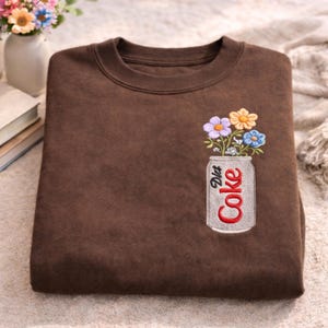 May include: A brown sweatshirt featuring an embroidered design of a Diet Coke can filled with colourful flowers. The can is silver with the words "Diet Coke" in red. The flowers are purple, yellow, and blue.