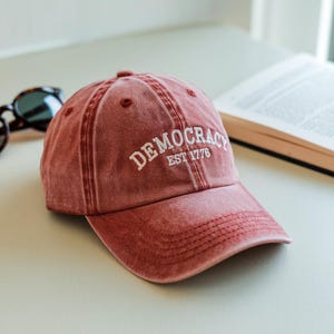 May include: A red baseball cap with the word "DEMOCRACY" embroidered in white, along with "EST 1776". The cap is made of a soft, faded material and has a curved brim. Sunglasses and a book are in the background.