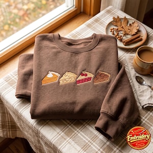 May include: A brown crewneck sweatshirt featuring an embroidered design of four distinct pie slices arranged in a horizontal row. The embroidery depicts a pumpkin pie slice with cream, an apple pie slice, a cherry pie slice, and a pecan pie slice.