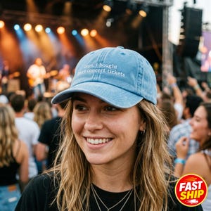 May include: A light blue baseball cap with the white embroidered text "oops, i bought more concert tickets" on the front panel. The cap features a curved brim, a structured crown, and a button on top, designed with a classic six-panel construction.