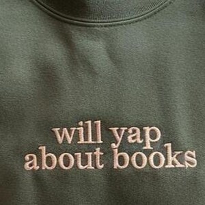 May include: Olive green sweatshirt with the embroidered text "will yap about books" in a light brown color. The crew neck sweatshirt is made of a soft material.
