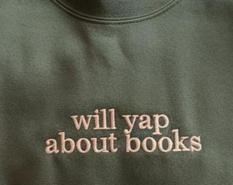 May include: Olive green sweatshirt with the embroidered text "will yap about books" in a light brown color. The crew neck sweatshirt is made of a soft material.