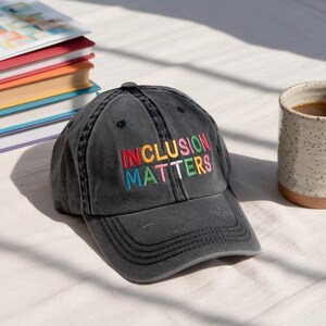 May include: A dark gray baseball cap with the embroidered text "INCLUSION MATTERS" in rainbow colors. The hat is sitting on a white surface, with a stack of books and a mug in the background.