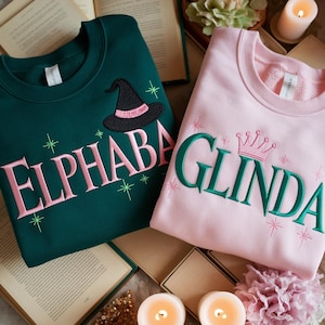 Pu&ograve; includere: Due felpe, una verde smeraldo e una rosa. La felpa verde presenta il nome "ELPHABA" in ricamo rosa, un cappello da strega e stelle. La felpa rosa ha "GLINDA" in ricamo verde, una corona e stelle.