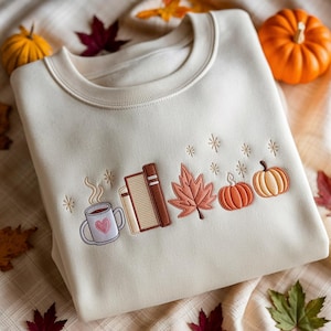Op de afbeelding: Crèmekleurige sweatshirt met een geborduurd herfstthema. Het ontwerp omvat een dampende mok met een hart, boeken, een esdoornblad, pompoenen en sneeuwvlokken. Twee kleine pompoenen en herfstbladeren zijn rond de sweatshirt verspreid.