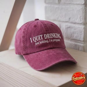 Peut inclure: Une casquette de baseball marron avec une finition délavée et usée. L'avant présente un texte brodé en blanc qui indique I QUIT DRINKING suivi du texte plus petit just kidding, i'm pregnant. La casquette a une visière courbée et des œillets.