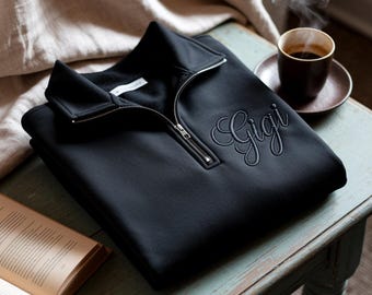 May include: A black sweatshirt with a quarter-zip neckline and the embroidered name "Gigi" in a cursive font. The sweatshirt is folded neatly on a light blue table, next to a cup of coffee and an open book.