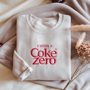 以下が含まれることがあります： 赤色の「I NEED A Coke Zero」の刺繍が施されたクリーム色のスウェットシャツ。スウェットシャツはクルーネックで、袖口が折り返された長袖です。柔らかくニュートラルな色調の表面に表示されています。
