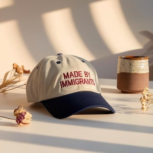 May include: A beige baseball cap with a navy blue brim and the embroidered text "MADE BY IMMIGRANTS" in red. A ceramic mug with a brown base and a speckled white top is to the right. Dried flowers are scattered around.