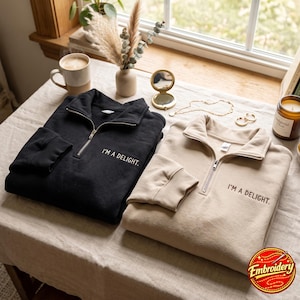 May include: Two quarter-zip sweatshirts, one black and one beige, each featuring the embroidered text "I'M A DELIGHT." on the left chest. Both garments have a high collar, a silver-toned metal zipper, and ribbed cuffs on the long sleeves.