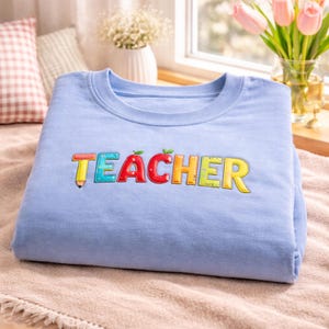 May include: A pale blue sweatshirt featuring the word "TEACHER" in a colourful embroidered design. The design incorporates a pencil, an apple, and various colours. The sweatshirt is neatly folded on a soft, neutral-toned surface.