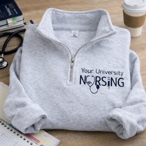 May include: A light gray quarter-zip sweatshirt with the text "Your University NURSING" in navy blue, along with a stethoscope and syringe graphic. The sweatshirt has a folded collar and a silver zipper. A coffee cup and medical supplies are in the background.