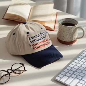 May include: A cream-colored baseball cap with a navy blue brim. The cap features embroidered text that reads "I want for you whatever you want for immigrants" in navy blue and red. A mug of coffee and an open book are also visible.